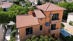 En bygning med flere etager, et rødt tegltag, en balkon med geometrisk design og udsigt til et bjergrigt landskab i baggrunden.