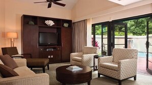 Una camera d'albergo con televisione a schermo piatto, ventilatore a soffitto, divano, poltrone, tavolino da caffè e vista su una piscina.