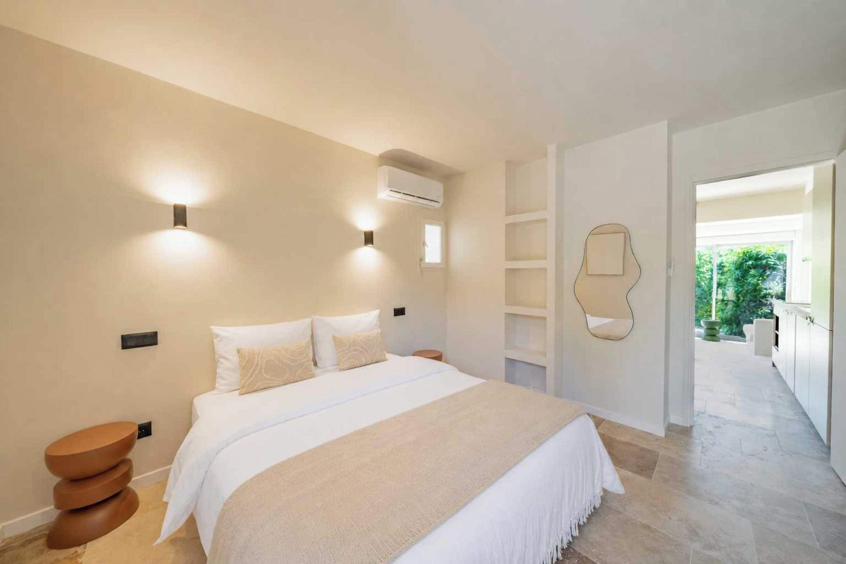 A bedroom with a bed, bedside tables, an air conditioning unit, and a view of the outdoors.