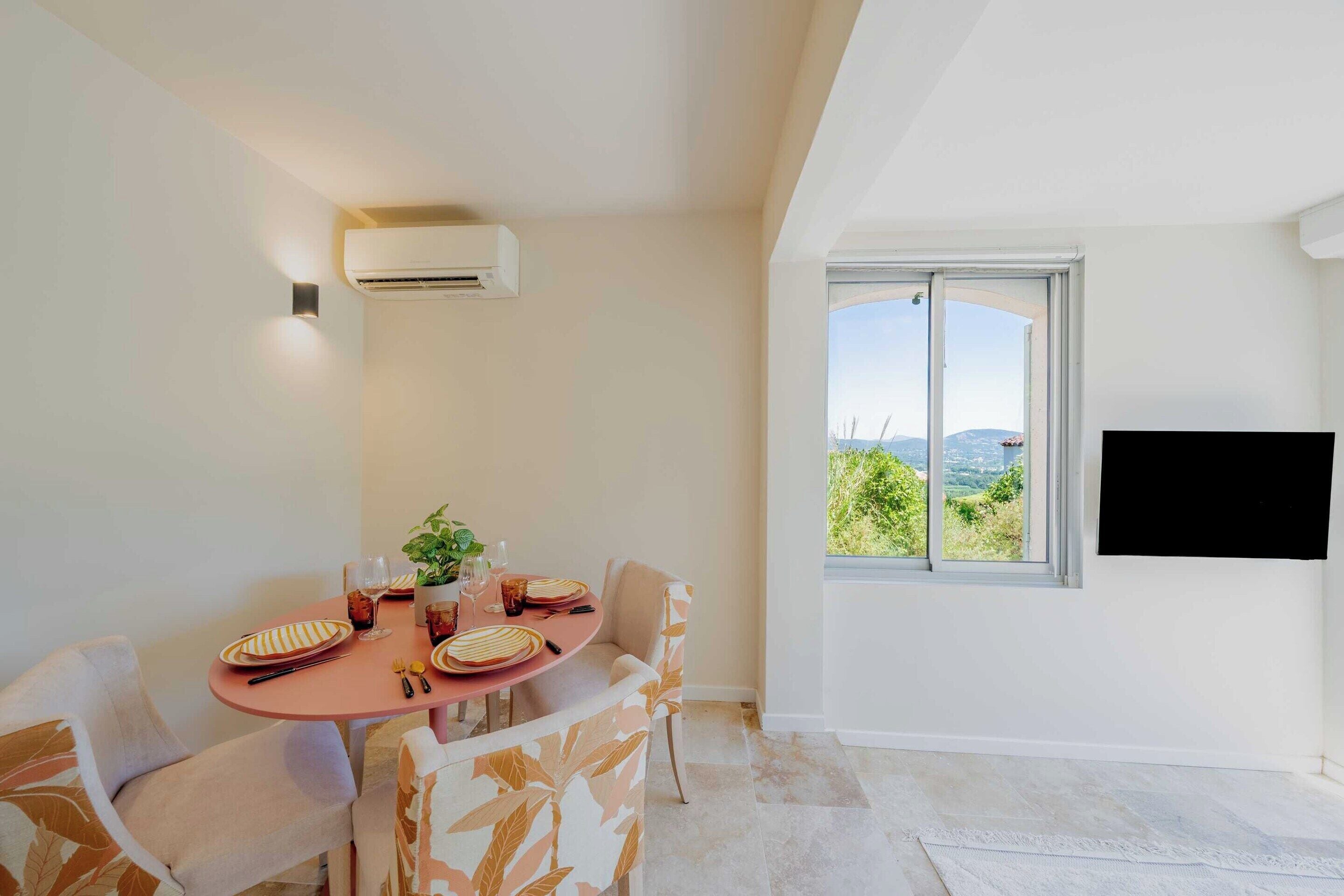 A dining area with a table set for four, a potted plant, and a large window offering a view of greenery and hills.