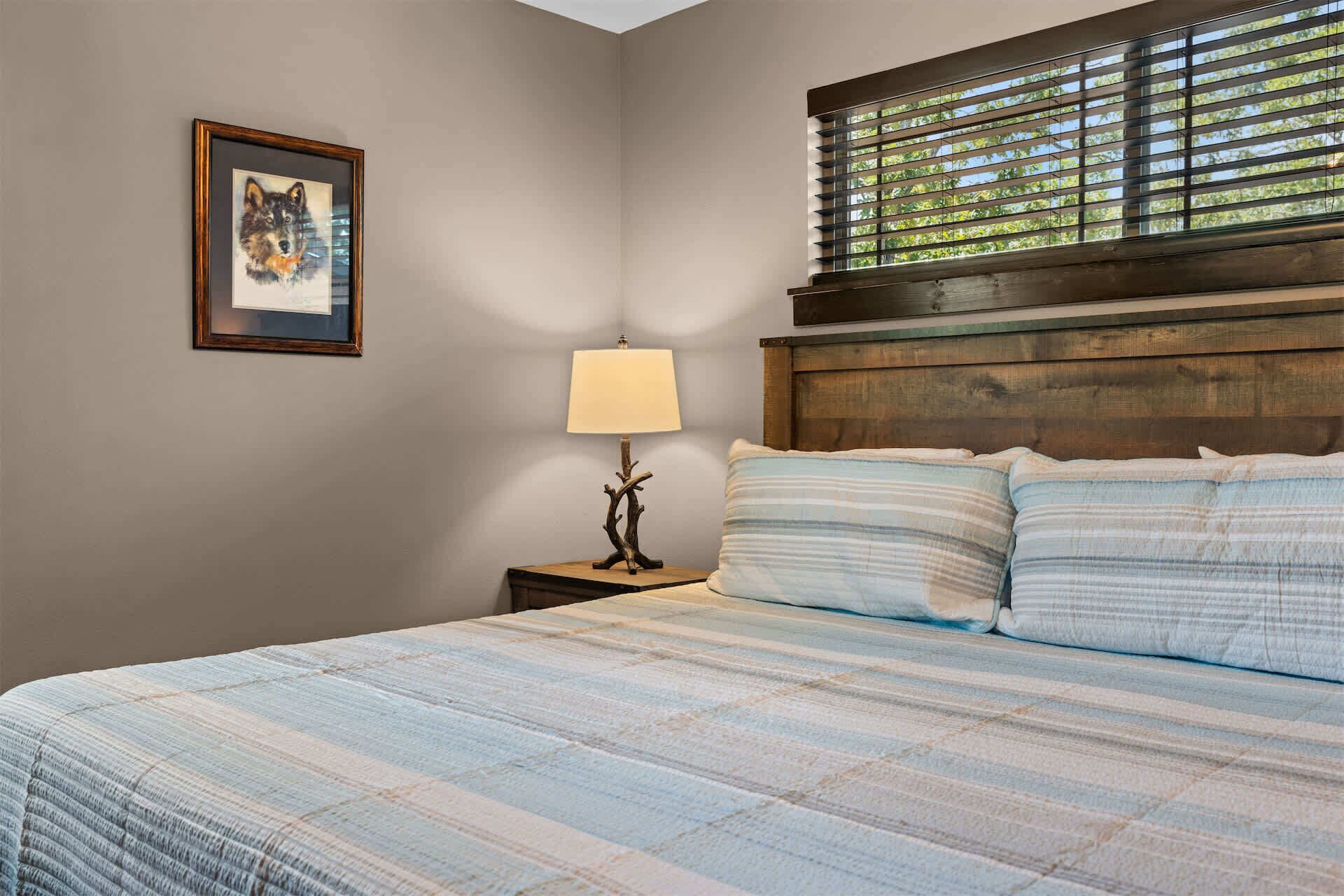 Une chambre à coucher avec un lit, une table de chevet, une lampe, un tableau encadré et une fenêtre avec des stores.