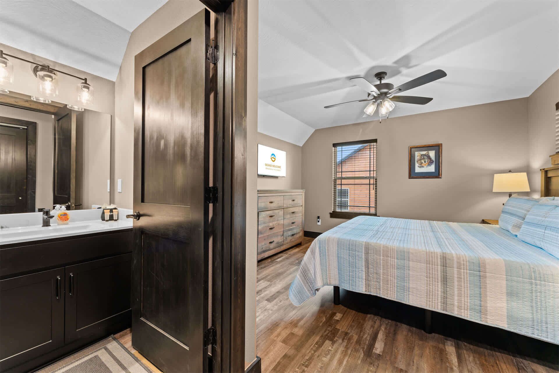 A bedroom with a bed, a ceiling fan, a window with blinds, a dresser, and a bathroom with a mirror and sink.