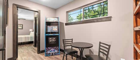 A room with a gaming console, a round table, two chairs, a window with a view of trees, and a door leading to another room.