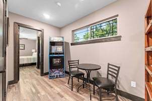 A room with a gaming console, a round table, two chairs, a window with a view of trees, and a door leading to another room.