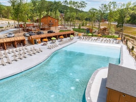A swimming pool with lounge chairs and umbrellas, surrounded by wooden buildings and a hilly landscape.