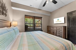 Un dormitorio con cama, ventilador de techo, ventana con persianas, televisor y cómoda de madera.