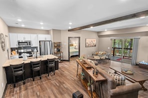 A modern kitchen with stainless steel appliances, a dining area with a wooden table and bar stools, and a large window with a view of the outdoors. - 8 Tall Oaks - Tall Timbers Escape (Hollister)