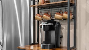 En kaffemaskin står på en hylla av trä, och ovanför hyllan finns ett metallställ där flera muggar hänger.
