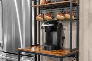 Una cafetera sobre un estante de madera con un escurreplatos metálico encima que sostiene varias tazas.