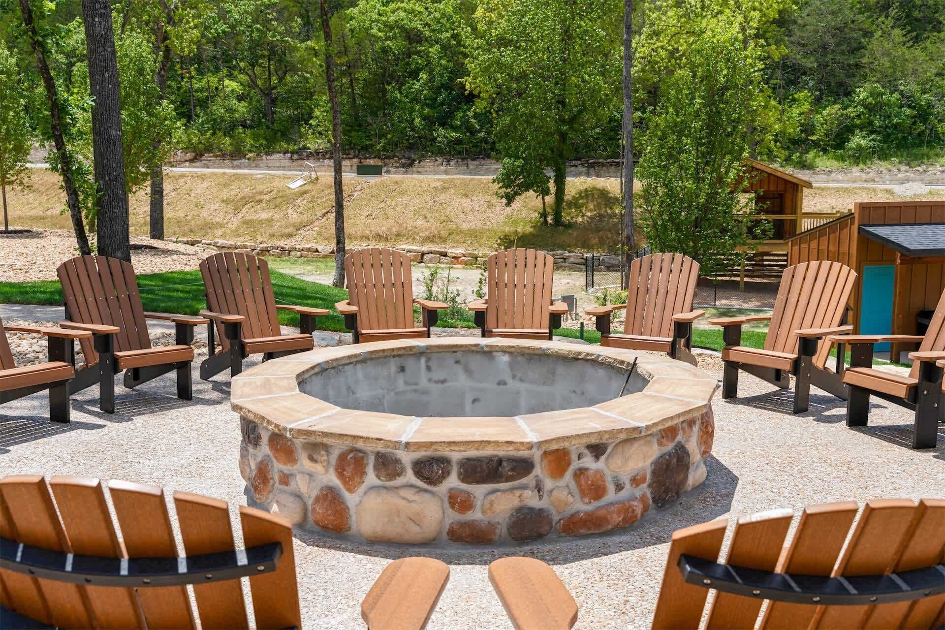 Un foyer circulaire en pierre, entouré de chaises Adirondack en bois, installé sur une surface de gravier avec des arbres en arrière-plan.