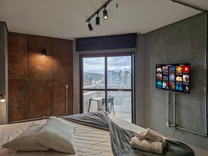 Una habitación de hotel moderna con una cama grande, vista de la ciudad a través de un ventanal amplio, televisor de pantalla plana en la pared y un balcón con una silla.