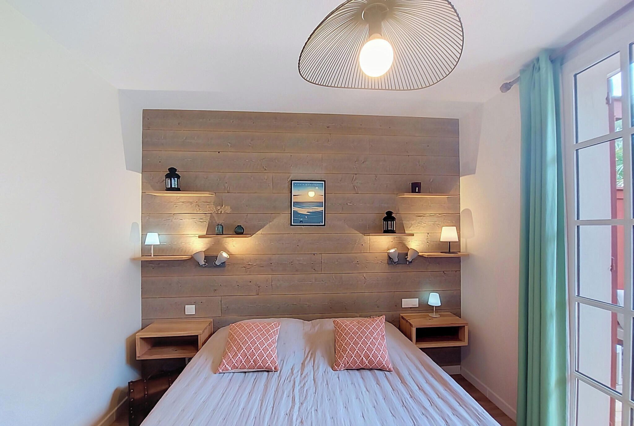 A bedroom with a wooden headboard, two bedside tables with lamps, a bed with patterned pillows, and a wall-mounted shelf with a framed picture.