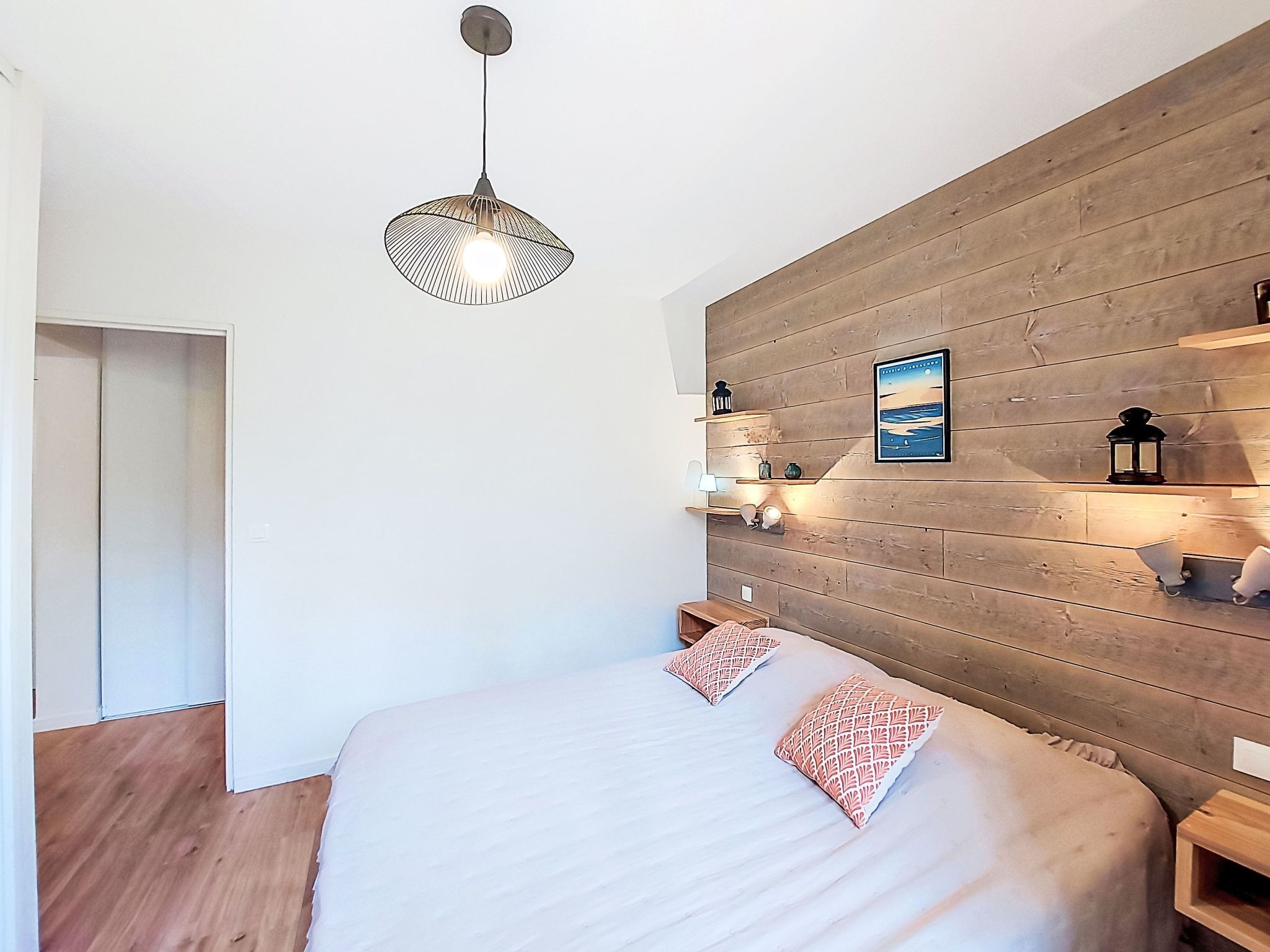 A bedroom with a wooden headboard, a bed with two pillows, a hanging light fixture, and a wall-mounted shelf with decorative items.