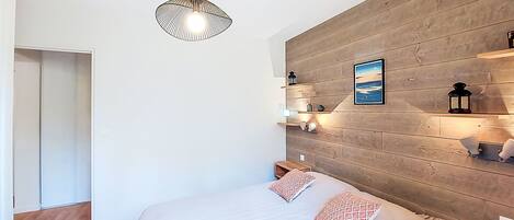 A bedroom with a wooden headboard, a bed with two pillows, a hanging light fixture, and a wall-mounted shelf with decorative items.