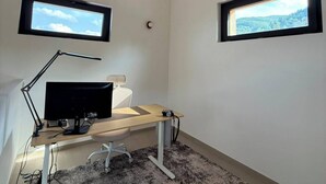 Ein Zimmer mit Schreibtisch, Computer und zwei Fenstern mit Aussicht.