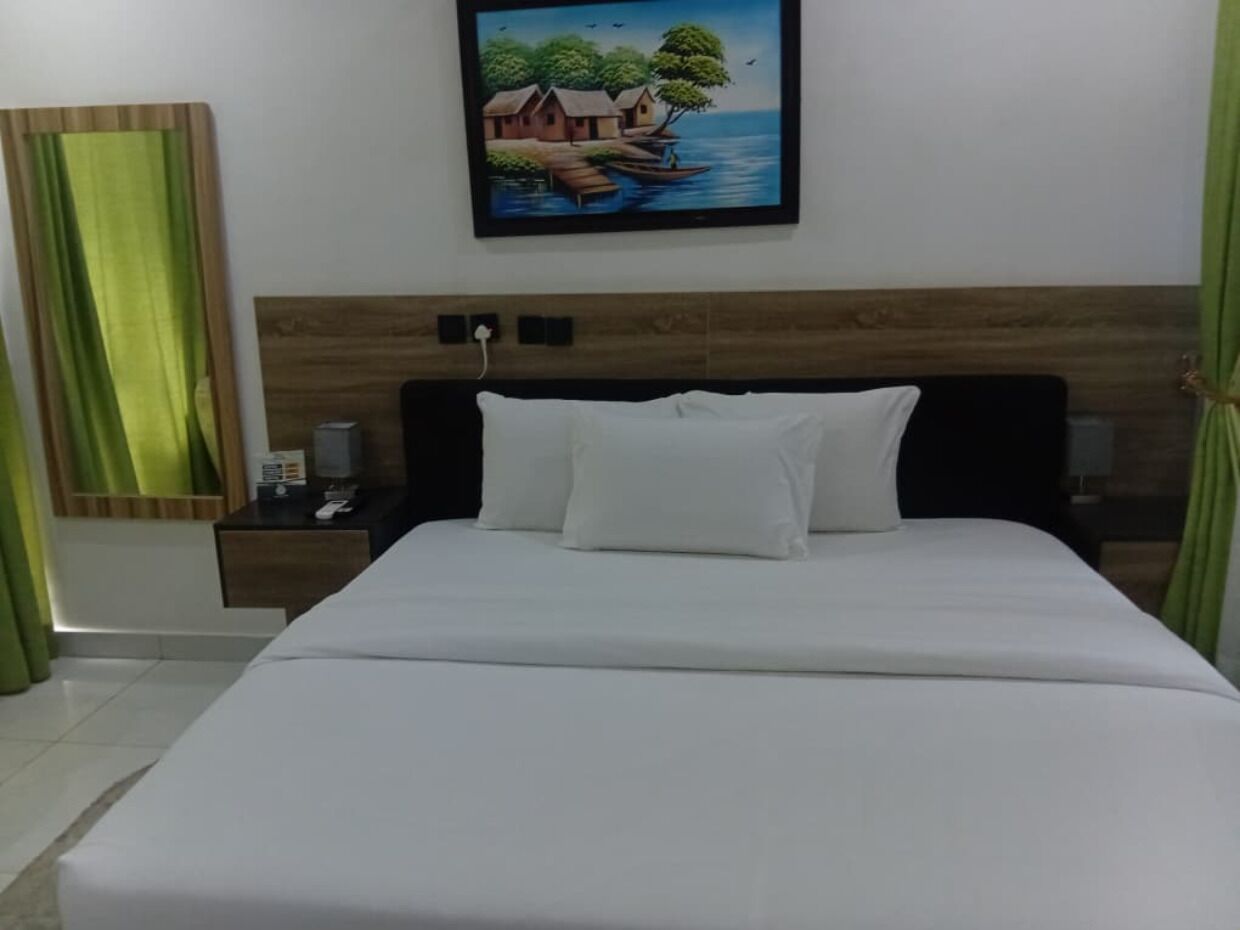 A bed with white bedding, a wooden headboard, a nightstand with a phone, and a painting of houses by a water body.