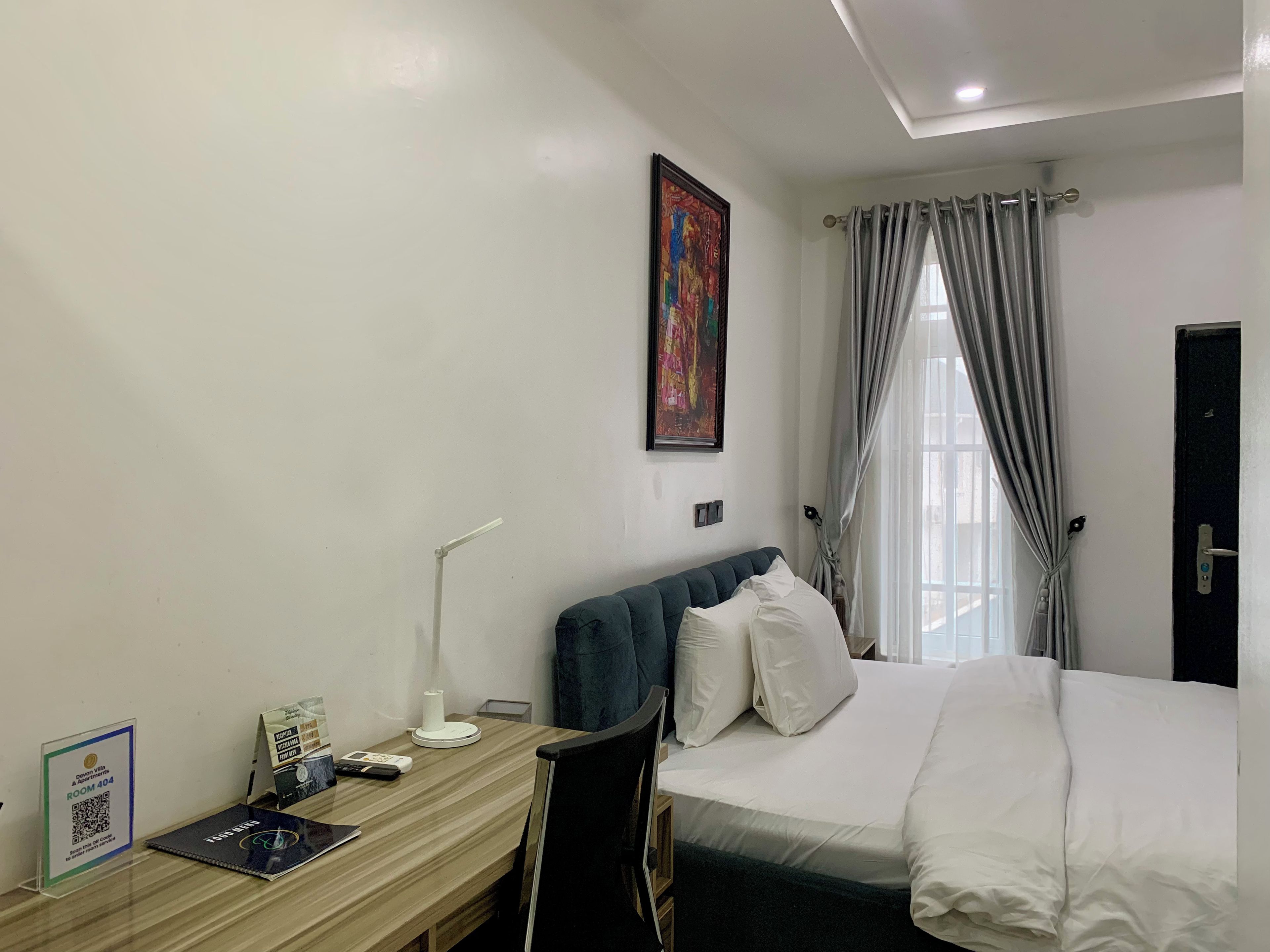 Een hotelkamer met een bed, een bureau met stoel, een bureaulamp, een lijst met foto aan de muur en een raam met gordijnen.