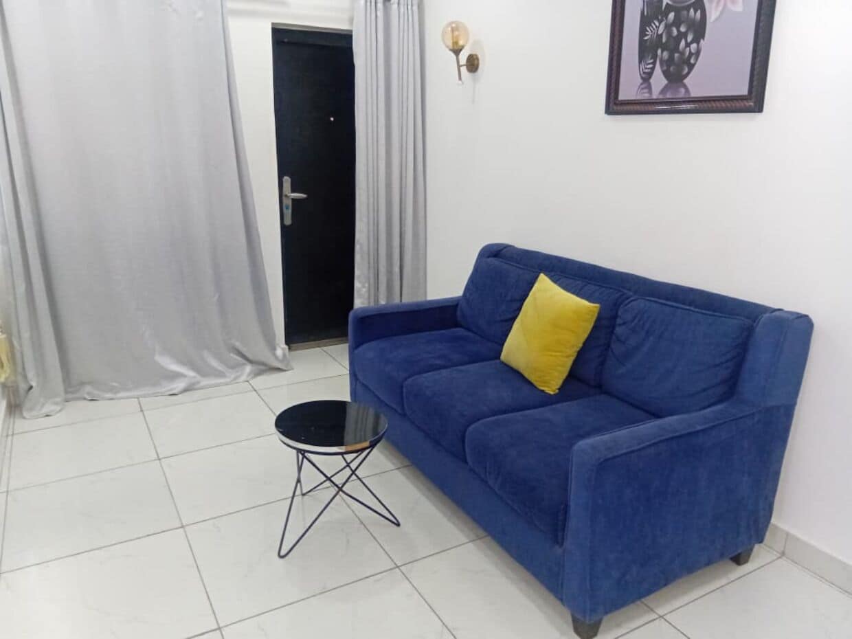 A blue sofa with a yellow pillow, a small black round table, and a door with a light switch on the wall.