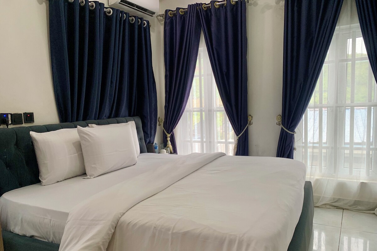 A neatly made bed with white bedding and pillows, a dark green headboard, and a window with blue curtains.