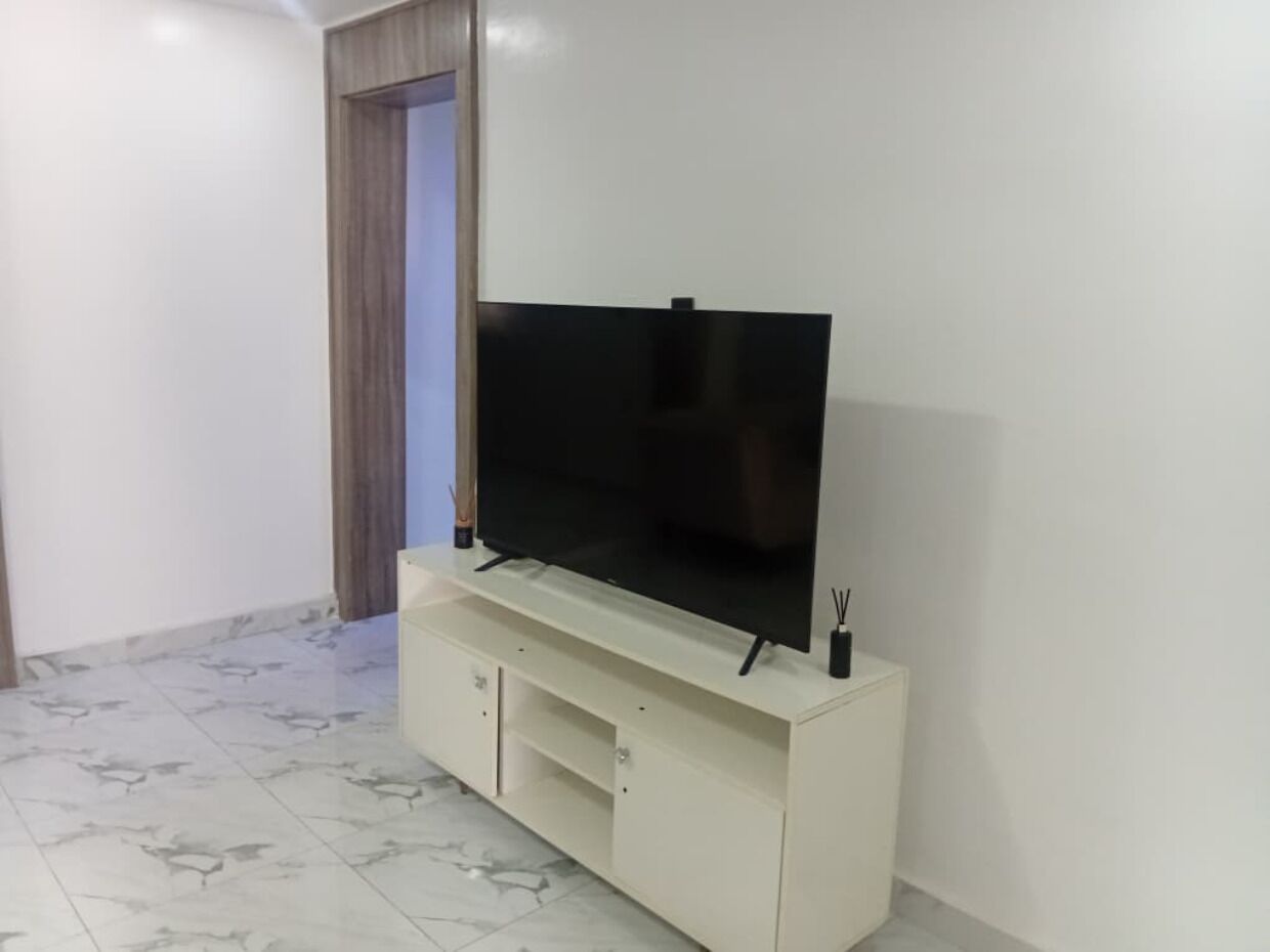 Een kamer met een flatscreen televisie op een witte kast, een deur met gordijnen, en een betegelde vloer.