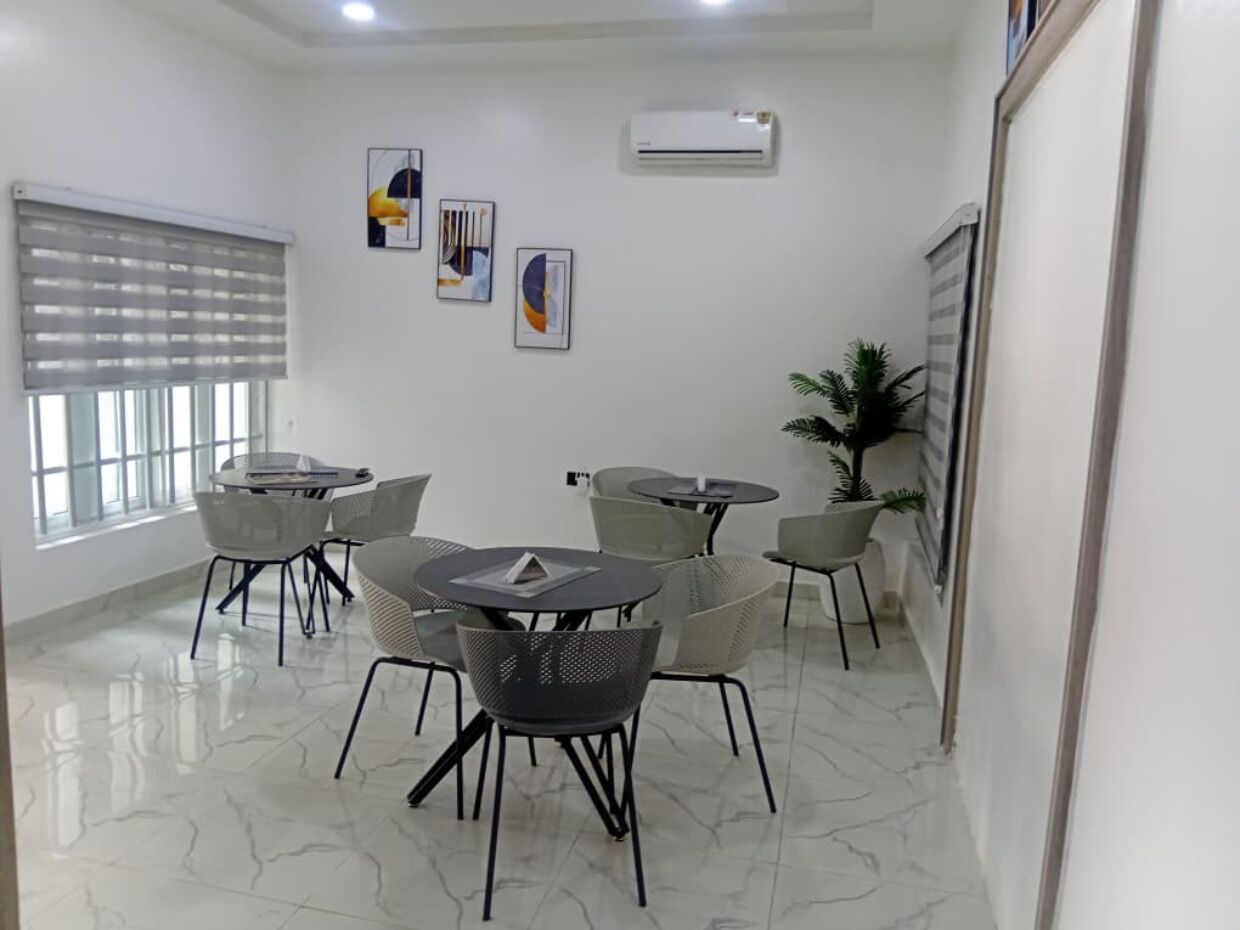 A modern conference room with white walls, a marble floor, and a large window with blinds. There are several round tables with chairs, and a potted plant is visible.