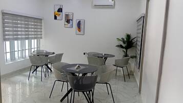 A modern conference room with white walls, a marble floor, and a large window with blinds. There are several round tables with chairs, and a potted plant is visible.