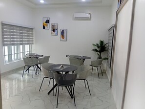 A modern conference room with white walls, a marble floor, and a large window with blinds. There are several round tables with chairs, and a potted plant is visible. - Devon's Villa and Apartments Apo (Abuja)