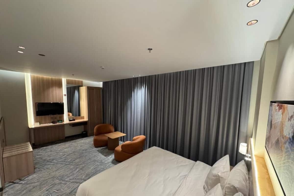 Une chambre d’hôtel dotée d’un grand lit, d’un bureau avec un téléviseur à écran plat, d’un coin salon avec des fauteuils et d’une grande fenêtre avec des rideaux.