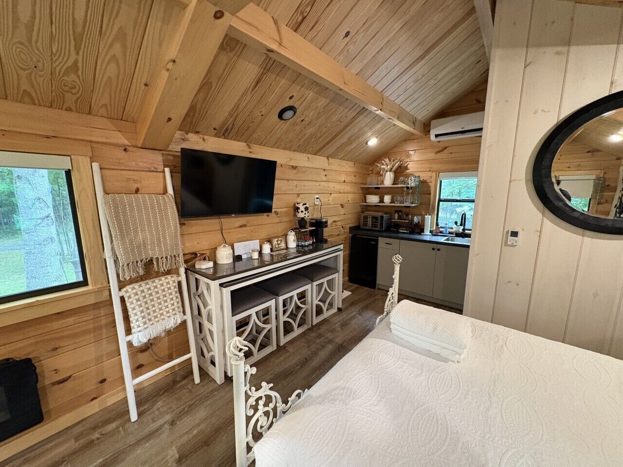 Interior acolhedor de uma cabana com cama, TV, cozinha compacta e uma grande janela.