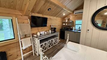 Interior acolhedor de uma cabana com cama, TV, cozinha compacta e uma grande janela.