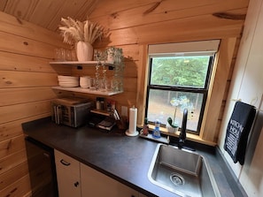 A compact kitchen with wooden walls, a window with a view of greenery, a sink, and basic appliances.