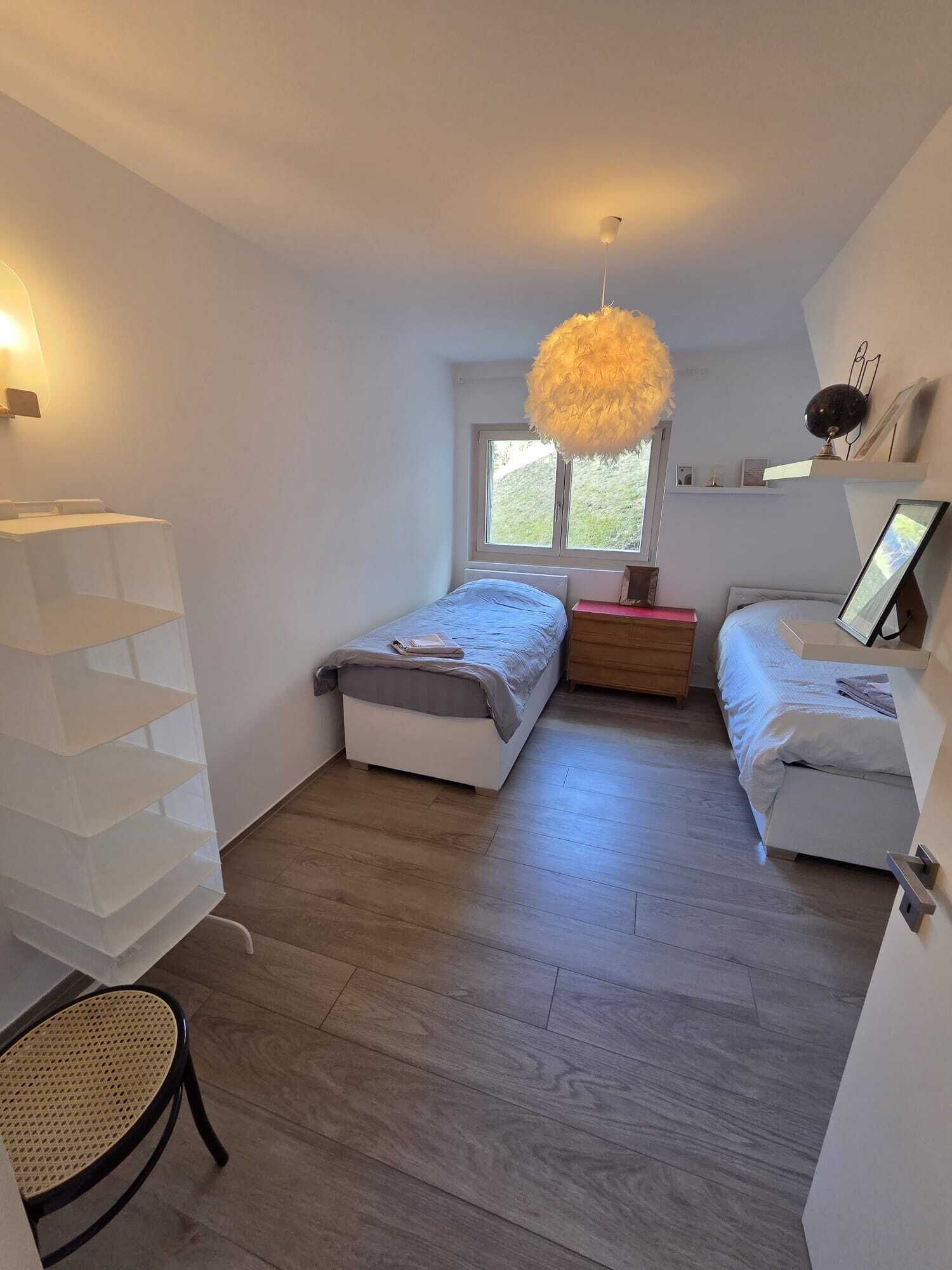 Ein Schlafzimmer mit zwei Betten, einem Fenster, einer Pendelleuchte und einem Rattan-Sessel.