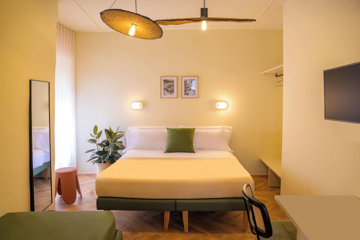 A modern hotel room with a large bed, bedside tables, a small stool, a potted plant, and a flat-screen TV.