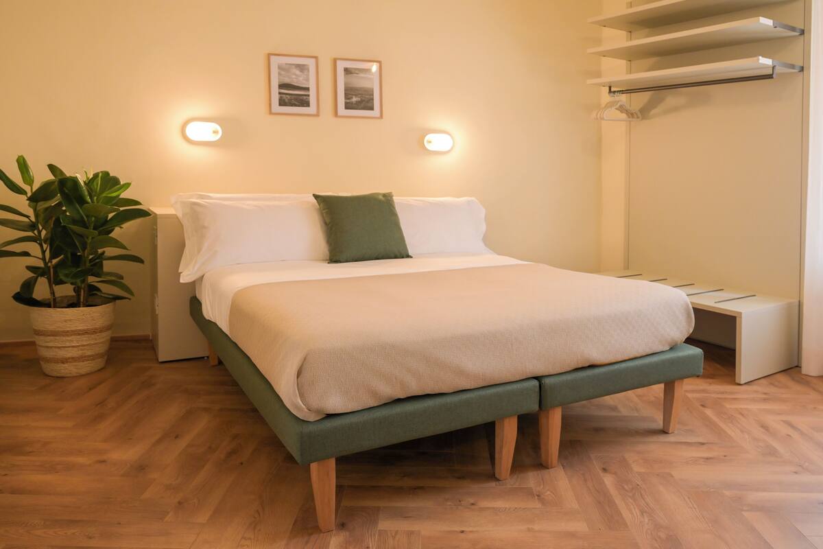 A neatly arranged bedroom with a bed, bedside tables, a potted plant, and wall-mounted lights.