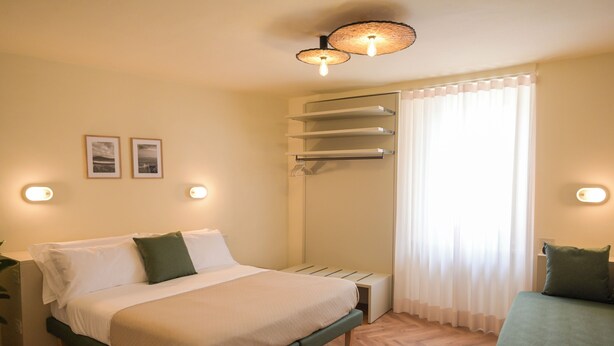 Une chambre à coucher comprenant un lit, des tables de chevet, un banc, une armoire et une fenêtre avec des rideaux.