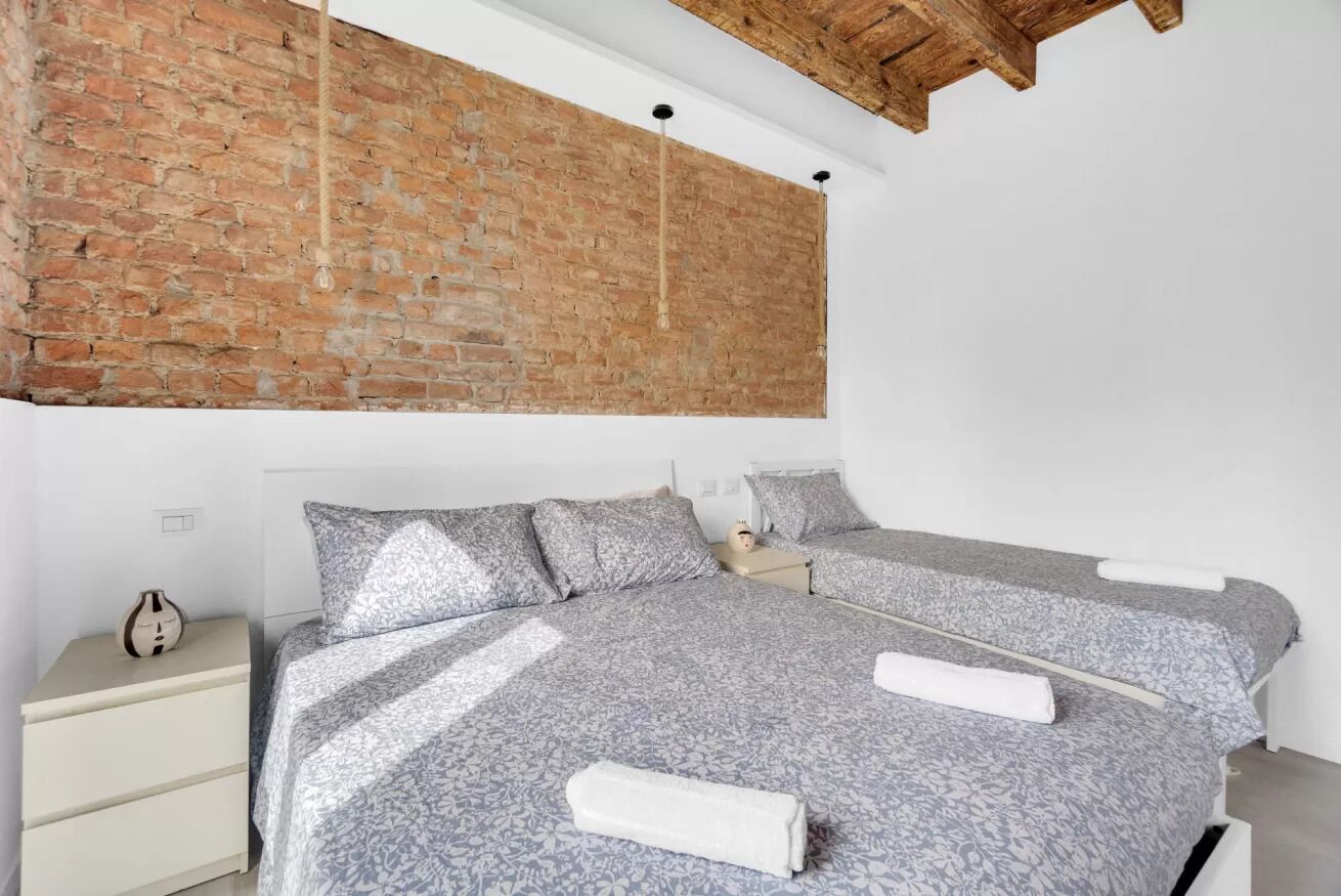 Una camera da letto con parete in mattoni, un letto con copriletto floreale grigio, un comodino e una lampada a sospensione.