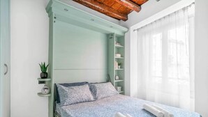 Ein Schlafzimmer mit einem Bett, einem Nachttisch, einem Regal mit einer Pflanze und einem Fenster mit durchsichtigen Vorhängen.