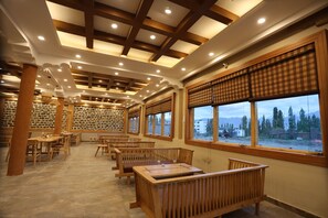 A spacious library with wooden tables and benches, large windows with blinds, and a stone wall. - The Grand View Ladakh (Leh)