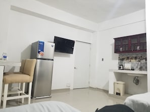 A compact hotel room with a kitchenette, a bed, a chair, and a TV mounted above the door.