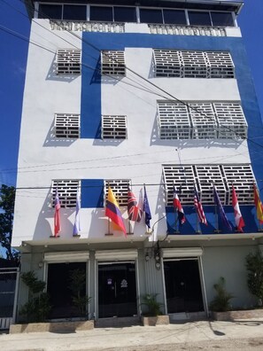 A multi-story building with a white facade and blue accents, featuring multiple windows with white grills and a row of flags at the ground level.