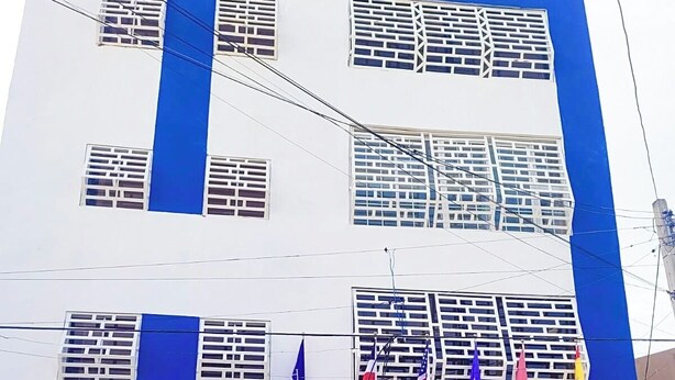 A multi-story building with a blue and white facade, multiple windows with metal grilles, and a row of flags at the entrance. - Yireh Hotel Grand Suites (San Juan de la Maguana)