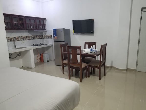 Habitación de hotel con cama, una mesa de comedor con sillas, zona de cocina con armarios y refrigerador, y un televisor de pantalla plana montado en la pared.