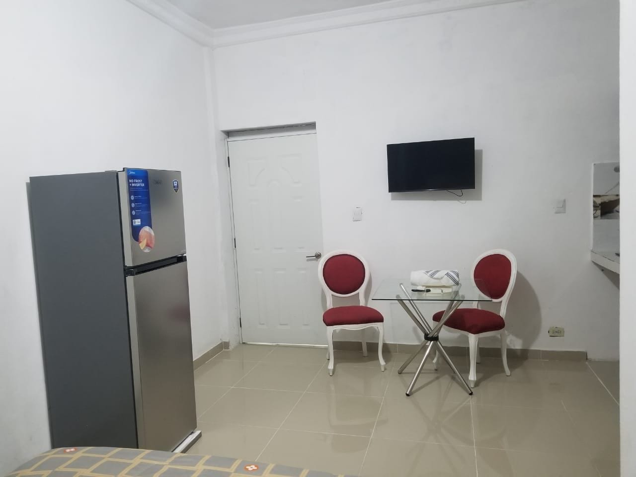 A compact kitchen with a refrigerator, a flat-screen TV mounted on the wall, and two red chairs around a small table.