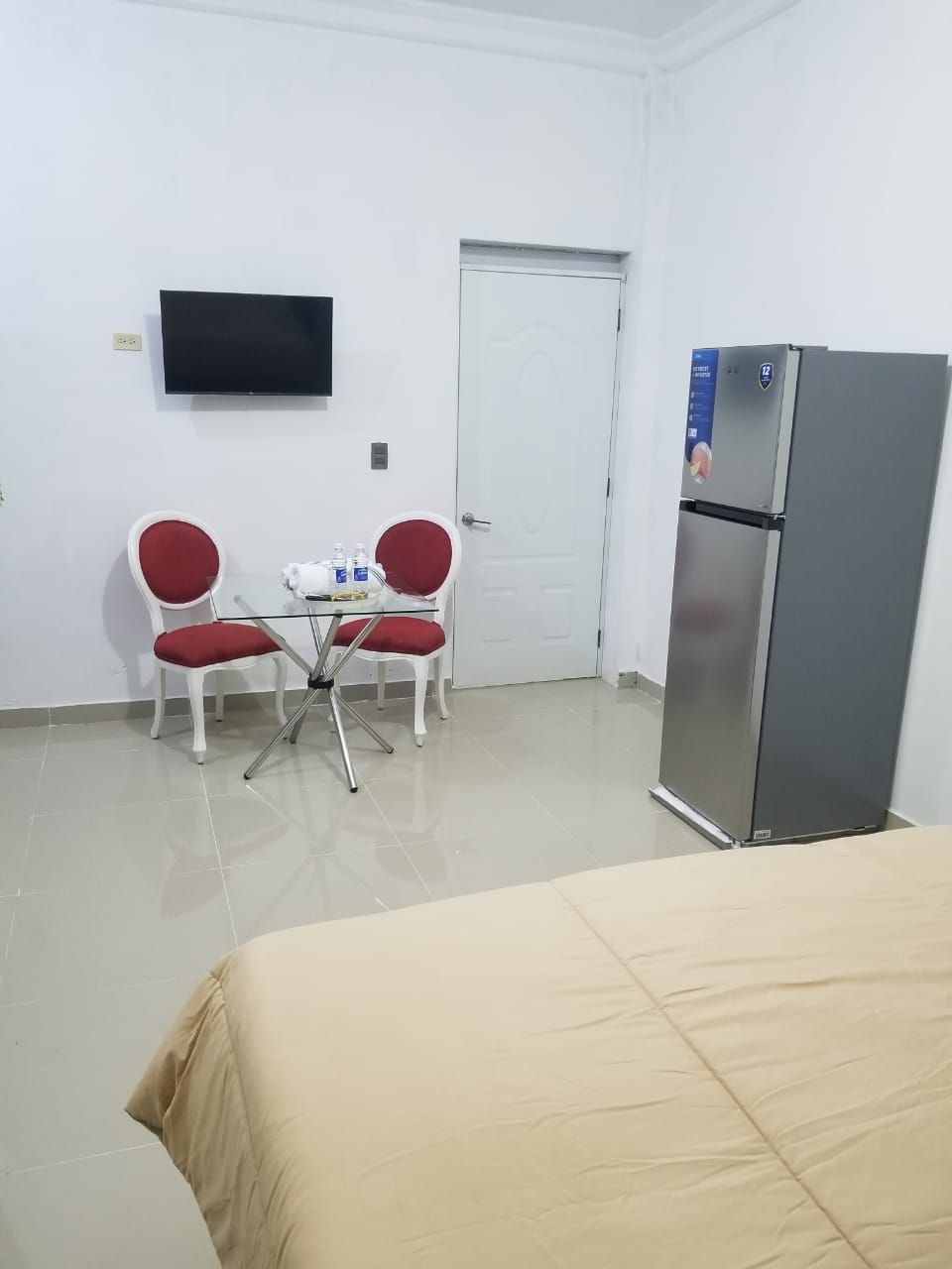 A hotel room with a bed, a television mounted on the wall, a refrigerator, and two red chairs around a small table with a teapot and cups.