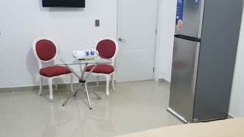 A hotel room with a bed, a television mounted on the wall, a refrigerator, and two red chairs around a small table with a teapot and cups.
