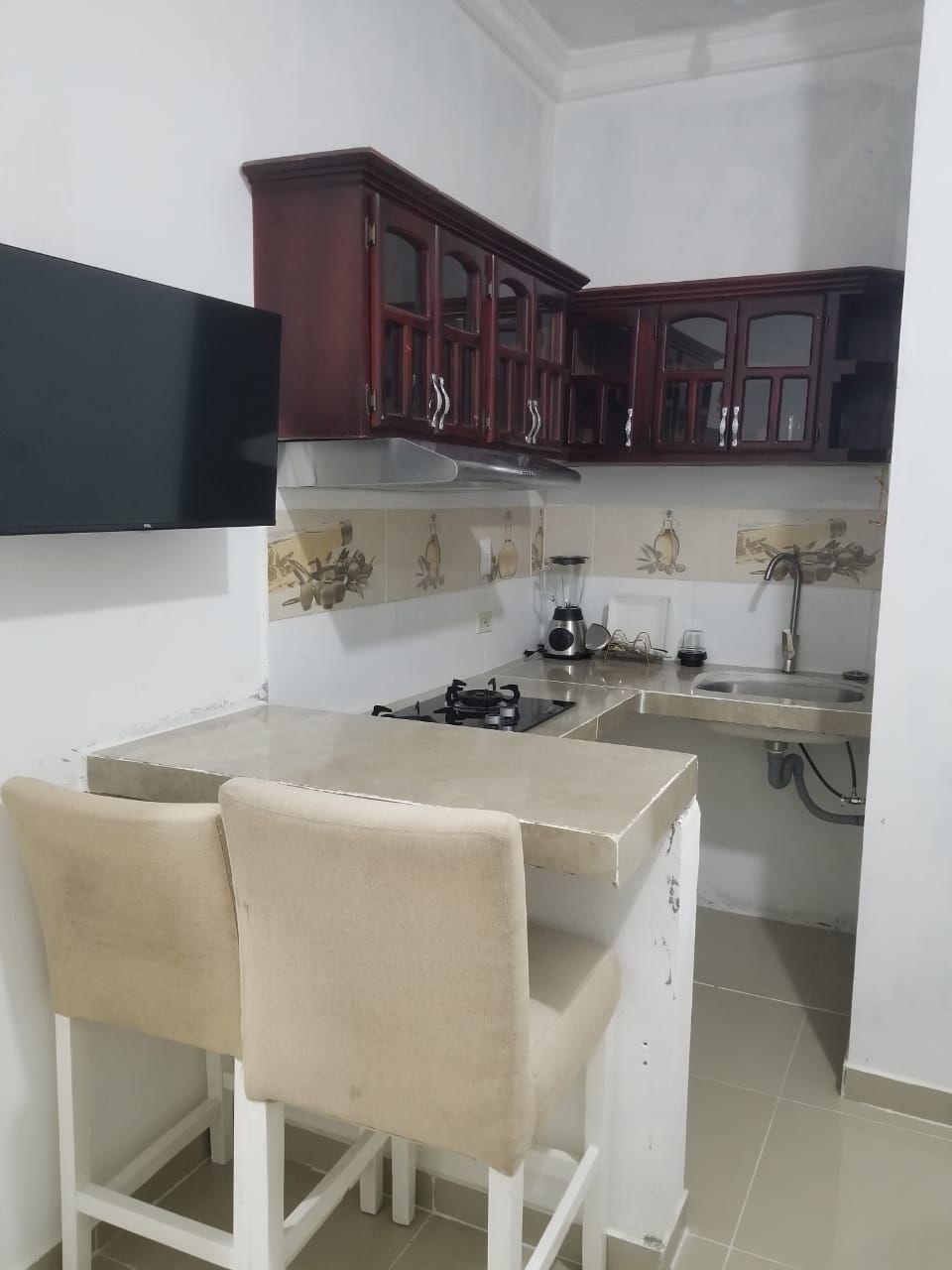 Eine kleine Küche mit Gaskochfeld, Spüle und einer Arbeitsplatte. Zwei Stühle und ein Schrank befinden sich über der Arbeitsfläche.