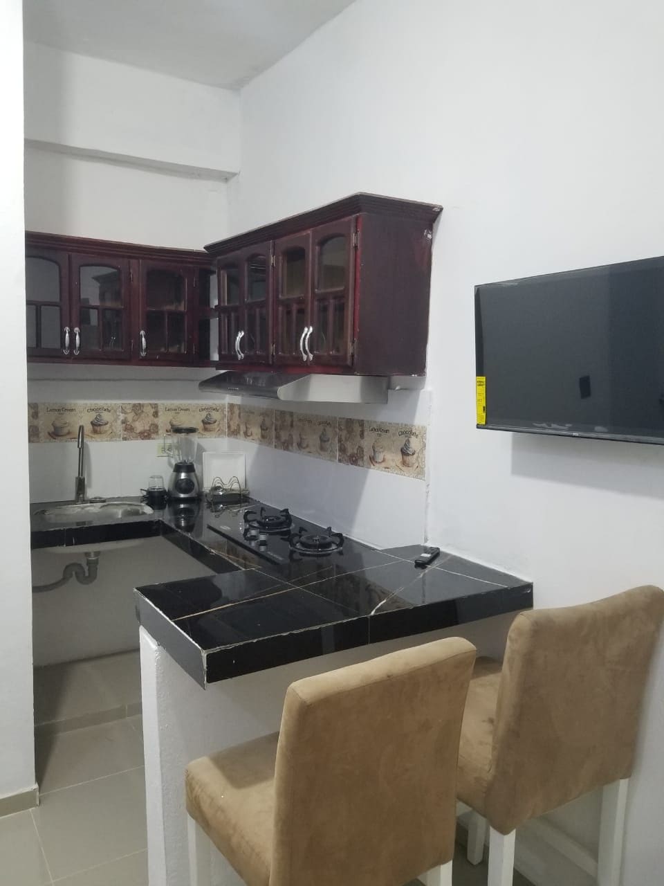 Una cucina con pensili in legno scuro, un piano cottura a gas, un piano di lavoro nero e due sedie beige.