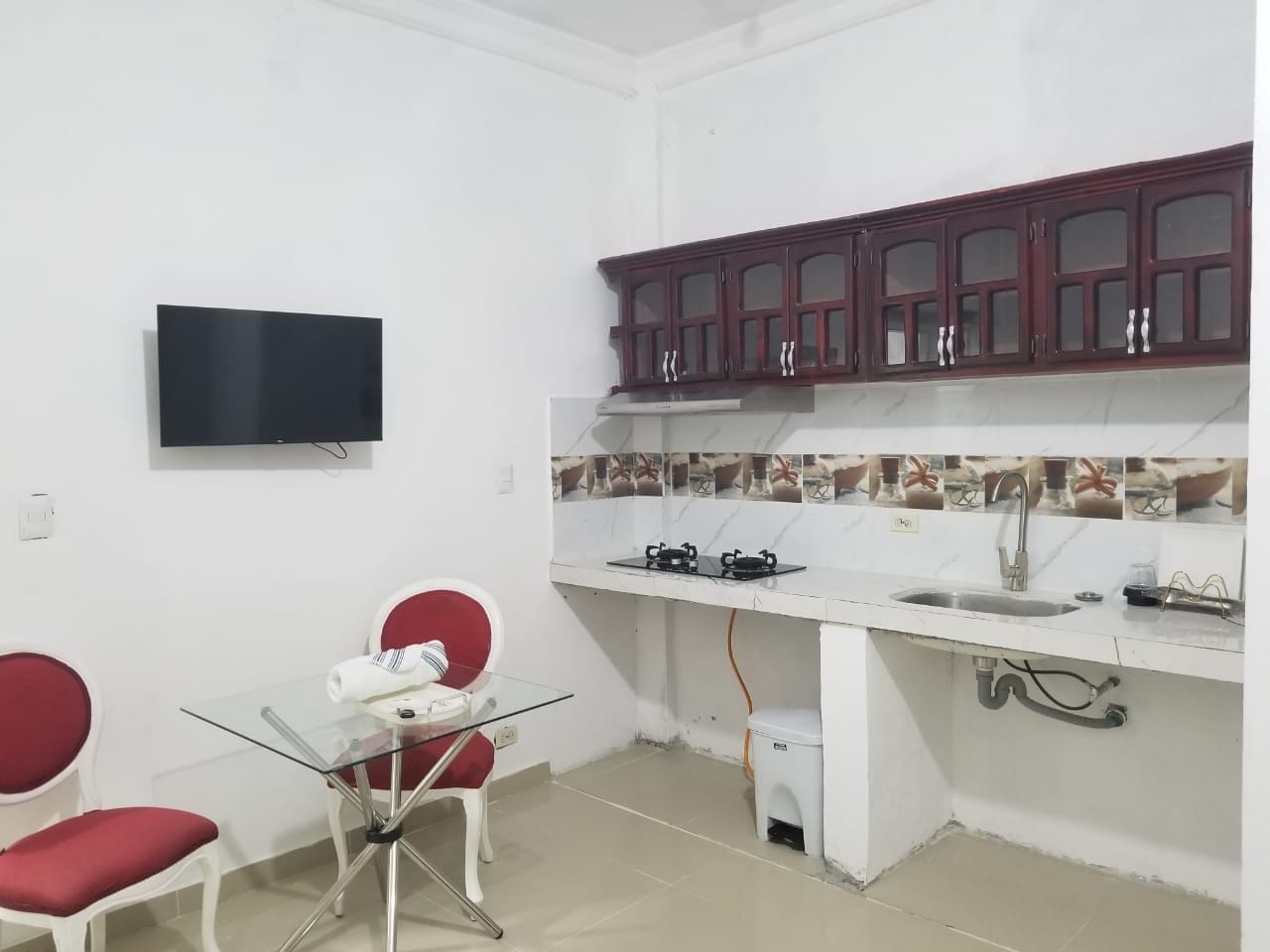 A compact kitchen with a TV, a dining area with two chairs, and a small trash bin.