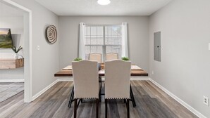 Un espace repas comprenant une table, des chaises, une fenêtre avec des stores, un luminaire au plafond et une prise électrique murale.