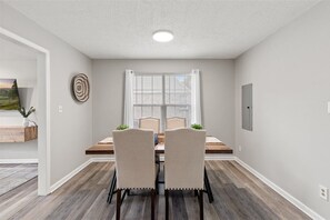 A dining area with a table, chairs, a window with blinds, a ceiling light, and a wall-mounted electrical outlet.